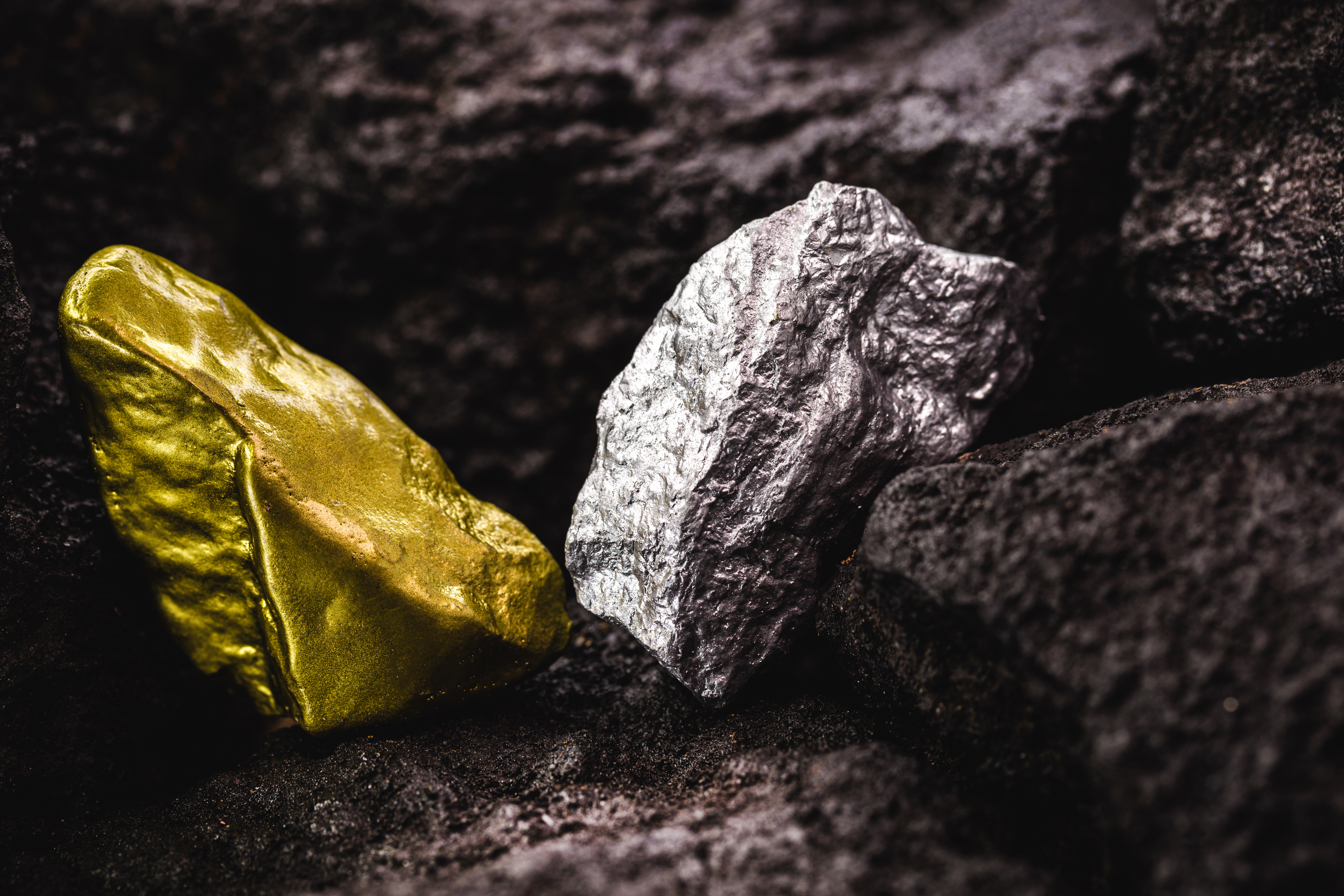 Raw gold and silver ore nuggets resting on dark, rocky ground.