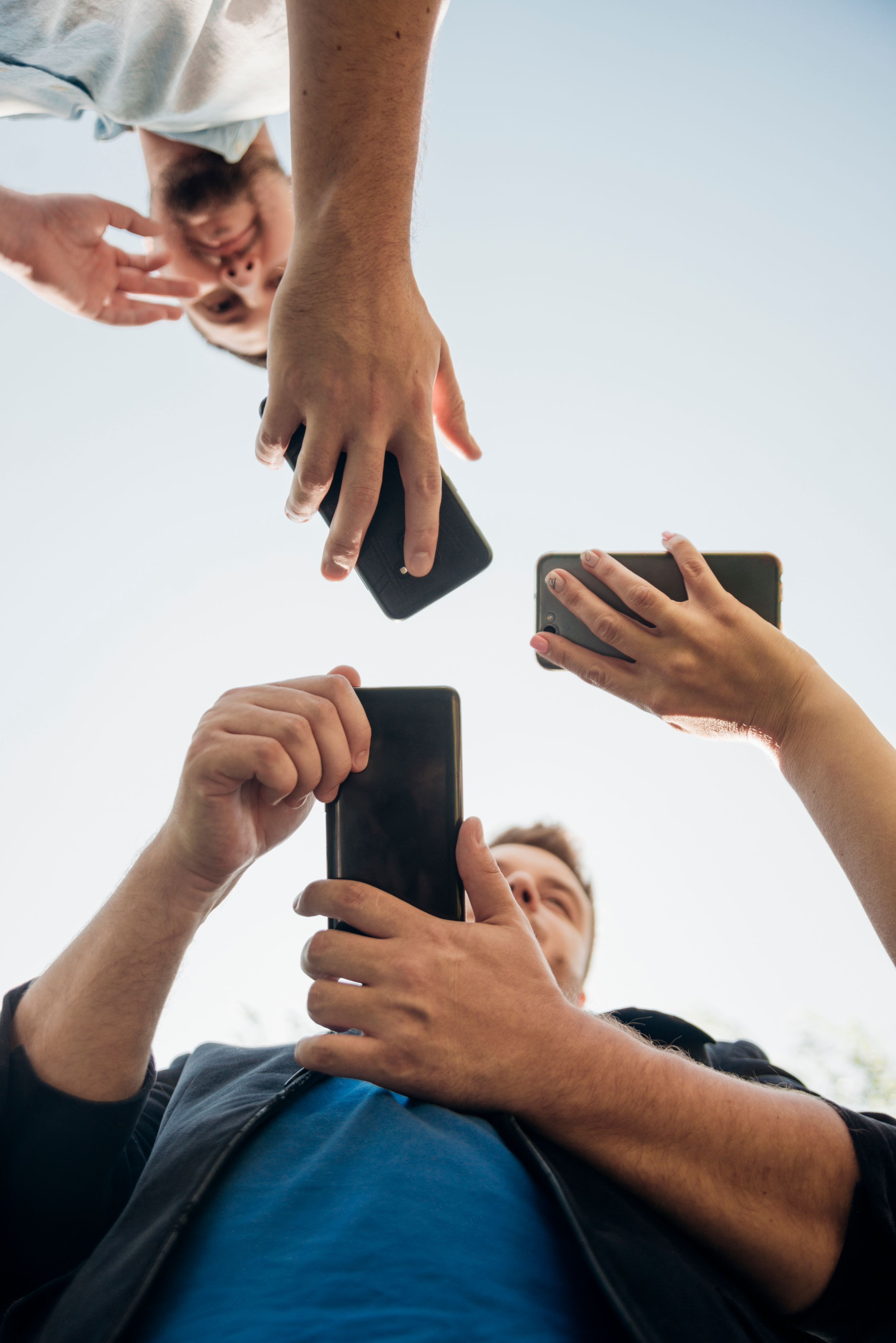 Low-angle view of three people looking down while holding smartphones against a bright, clear sky.